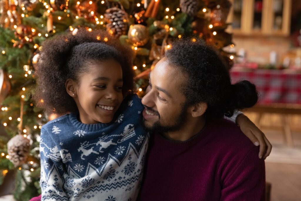 A father and daughter share a warm hug by the Christmas tree, enjoying a quiet moment together.